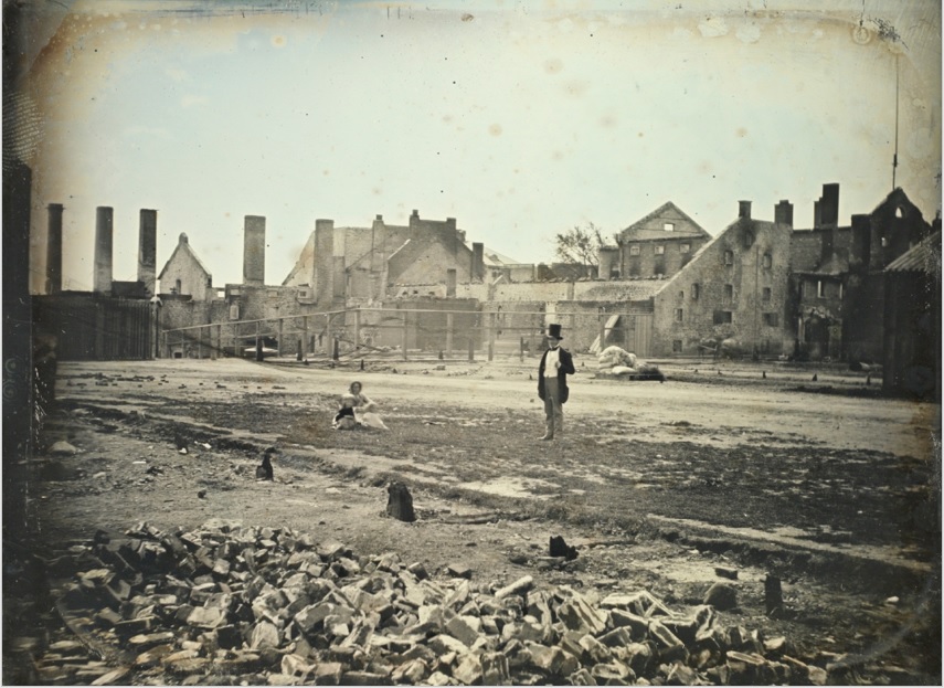 Chapter 2: Condition Issues. Image of a landscape image of a burnt building. A man in a top hat in the centre and a woman sitting on the ground off to the left. This photograph has a finger mark in the top left corner.