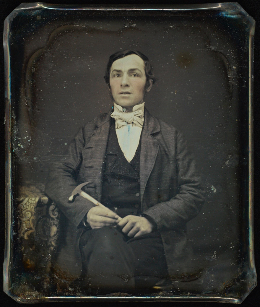 A hand-tinted daguerreotype portrait of a seated man holding a hammer.