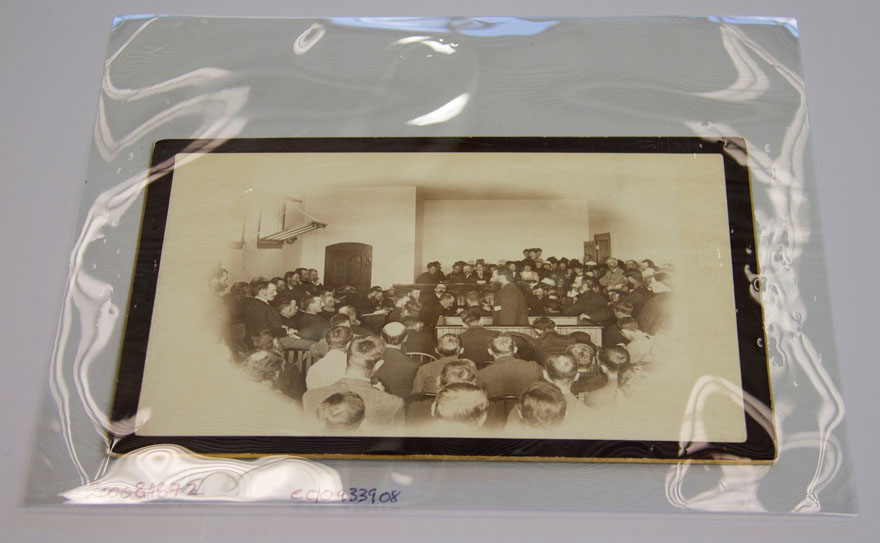 Image d'une photo rangée dans une enveloppe de plastique. Sur la photo, un homme est debout au milieu d'une salle. Il est entouré d'hommes assis qui le regardent, dans un tribunal.