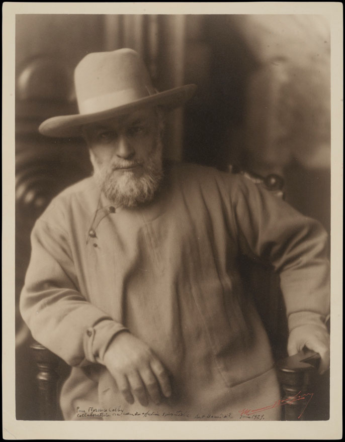 Man with a beard and a hat with a large rim, half seated, leaning on a chair arm.