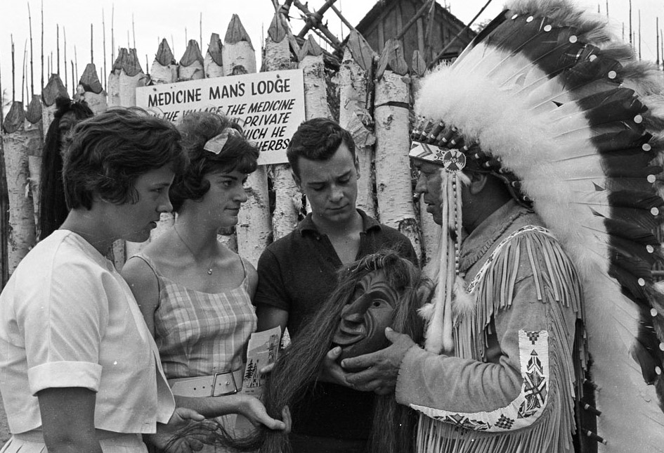 Black and white photo showing a log lodge in the background with an indigenous chief on the right holding a mask and speaking with 2 caucasian ladies and man on the left