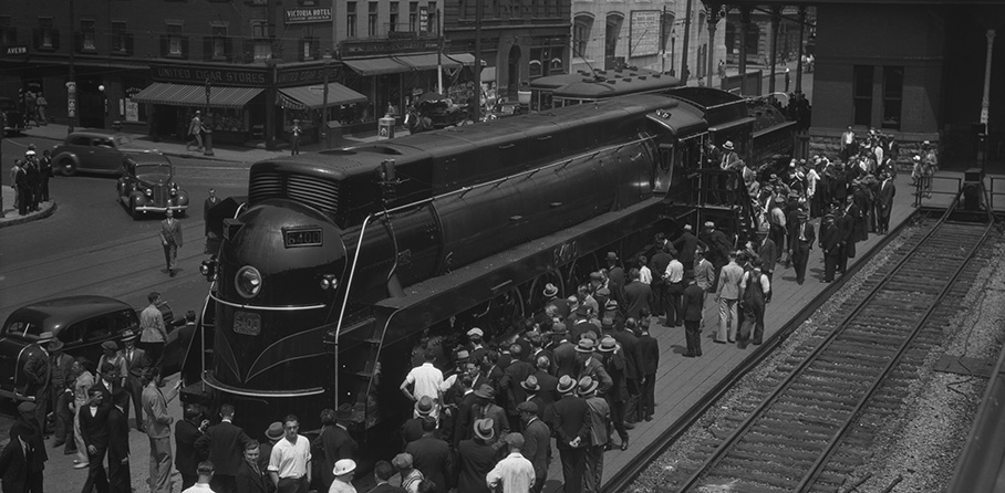 Photo en noir et blanc montrant une locomotive profilée, des passagers qui embarquent, un rail terminal à droite, et des édifices en arrière-plan.
