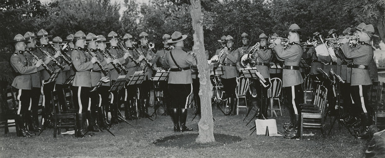 Photo en noir et blanc montrant des musiciens debout en uniformes de polices montée jouant des anches à gauche, des cuivres à droite avec le chef d'orchestre au centre et des arbres derrière
