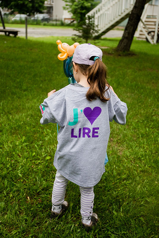 Photo couleur montrant une fillette vue de dos marchant sur le gazon, jouant avec une fleur en ballon et portant un gilet où il est écrit «J'aime lire»