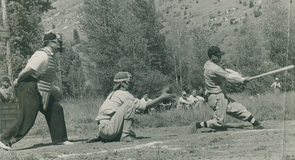 Photo noir et blanc montrant frappeur, receveur et arbitre de baseball avec spectateurs, arbres, et flanc de montagne en arrière-plan