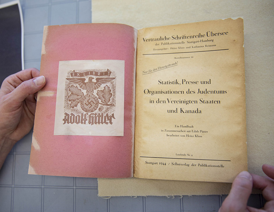 Photo de 2 mains tenant un livre ouvert montrant des symboles nazis et le nom «Adolf Hitler»  à gauche et l’éditeur, le titre, l’auteur et l’année (1944) à droite