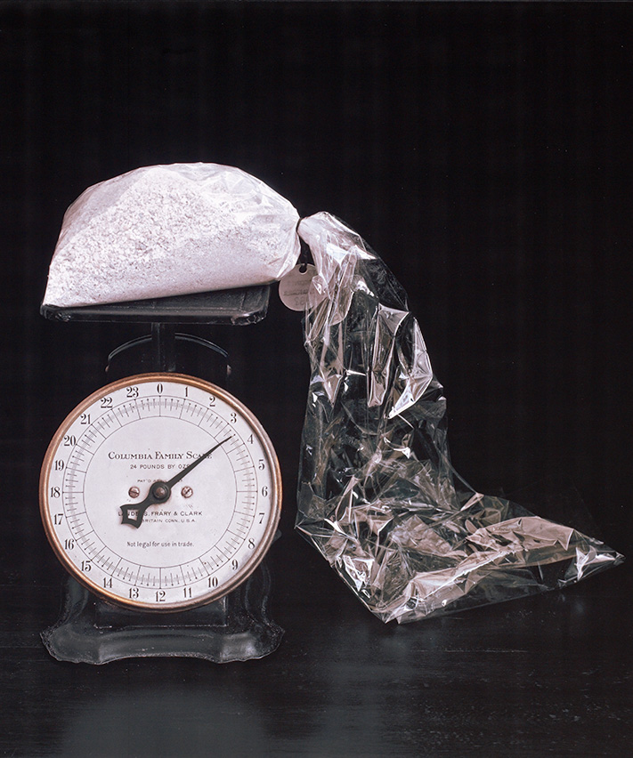 Color photo showing an old analog dial scale with dial, weighing a clear labeled bag filled with white powder