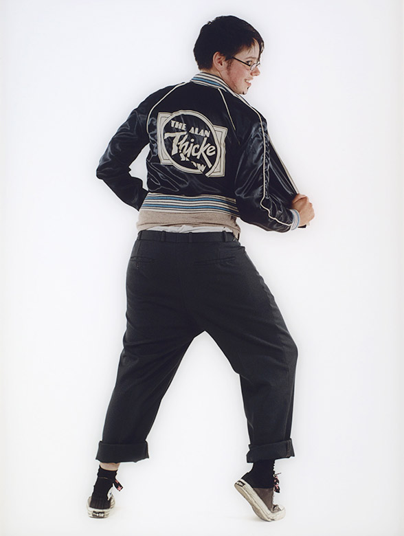 Color photo of a young man with glasses and goatee seen from the back, wearing a sports jacket , wide pants with cuffs, and sneakers