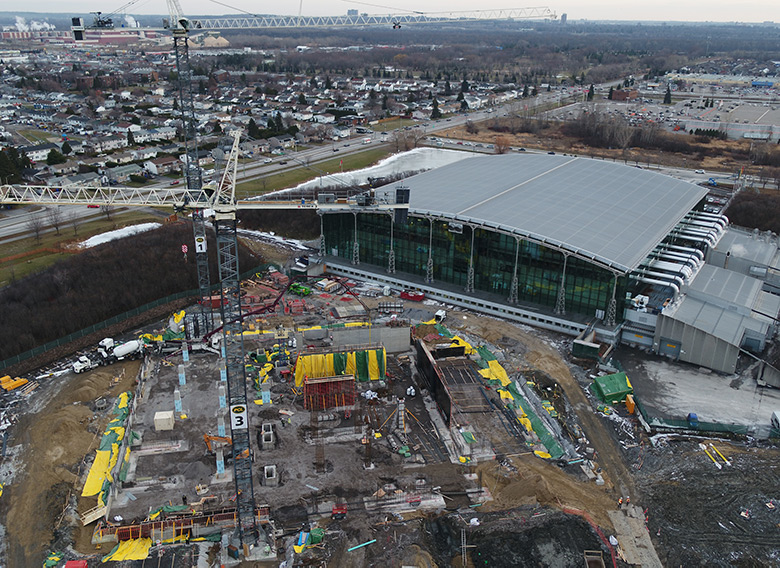 Photo couleur d’une vue à vol d’oiseau d’un site de construction montrant deux grues, une structure de fondation à l’avant-plan, et l’édifice Gatineau 1 et panorama semi-urbain en arrière-plan