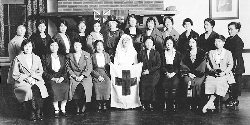Black & white photo showing 2 rows of predominantly Japanese women including one seated in the center front wearing a white dress and headdress holding a flag with a cross on it