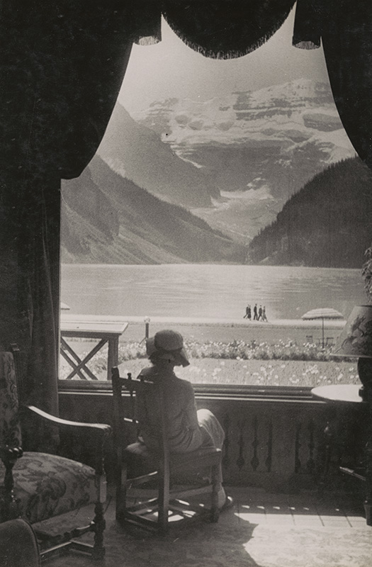 Black and white photo showing a seated woman seen from behind gazing from a huge window adorned with a swagged curtain, a mountainous panorama reflecting on a body of water