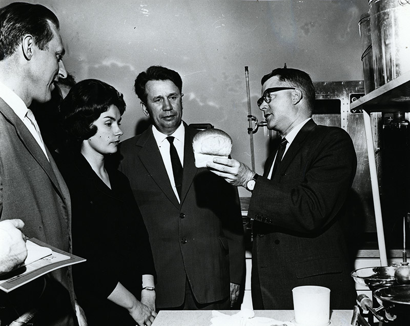 Photo noir et blanc d'une scène composée de poêlons et chaudrons à droite, suivi de 4 individus biens vêtus dont un homme tenant une miche de pain qu'observe 2 hommes et 1 femme à gauche