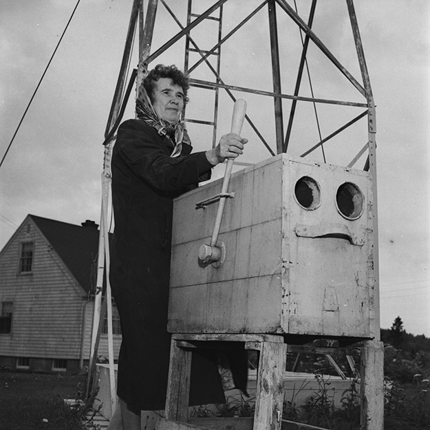 Black and white photo showing a small house and a metal tower behind a woman dressed in a veil and coat pressing a handle connected to a box with 2 openings on the front