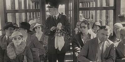 Black and white photo of a daytime scene showing a windowed train cabin with a porter capped with a kepi holding a large handkerchief and several pairs of sunglasses, walking between 2 rows of passengers