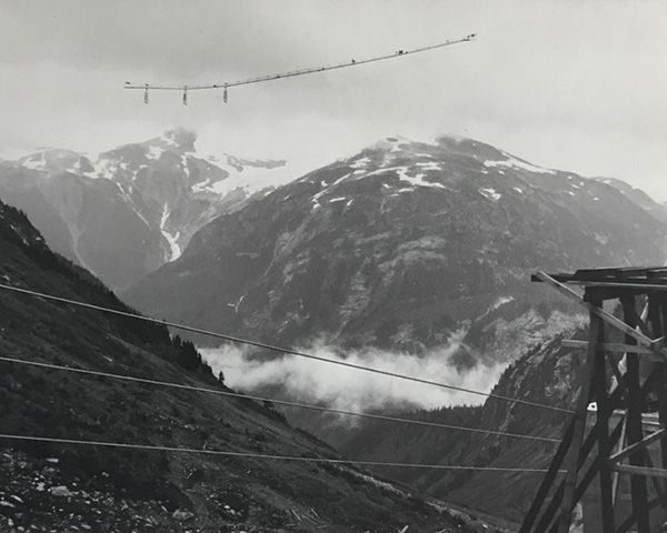 Black & white photo showing 2 snow capped mountains fading in the background, a mountain side, suspended power lines and a wooden structure in the foreground