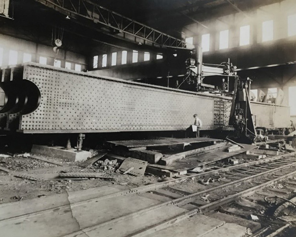 Black & white photo showing a high ceiling room with a sunlit window strip on top background, a pulley mechanism, 3 large steel beams and a worker, and rail tracks at the bottom foreground