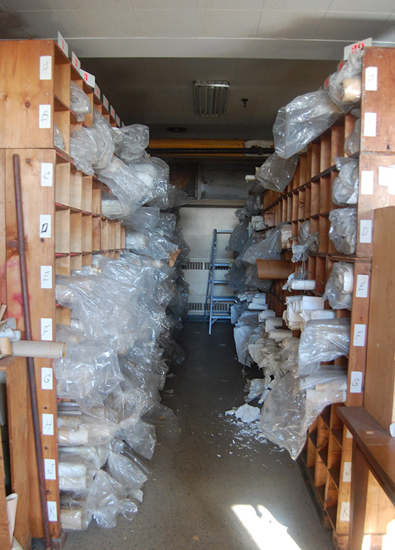 Color photo of a small and dim warehouse showing 2 rows of compartmented shelves filled with paper rolls in transparent bags, seperated by a narrow aisle in the middle.