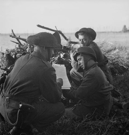 Black and white photo showing a field in the background, a mound, and 3 soldiers (one crouching holding a map, one sitted, and another kneeling holding a  submachine gun) in the foreground