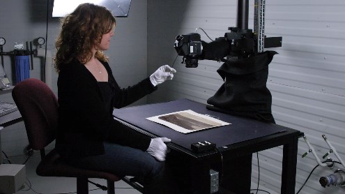 color photo of a small dark room showing a woman wearing white gloves sitting in front of a small table and photographing a monochrome document