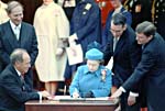 Colour photograph of Queen Elizabeth II seated at a table signing a document while others, including Prime Minister Trudeau, look on