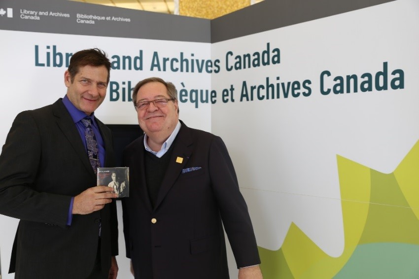 Guy Berthiaume and Micah Barnes standing in front of a banner with the words Library and Archives Canada. Micah Barnes is holding his CD. 