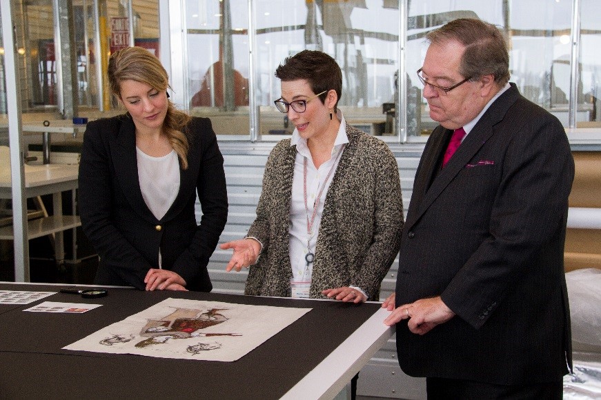 L’honorable Mélanie Joly, ministre du Patrimoine canadien, et Guy Berthiaume, bibliothécaire et archiviste du Canada, écoutent les explications de l’archiviste en art Geneviève Morin concernant le dessin d’un costume.